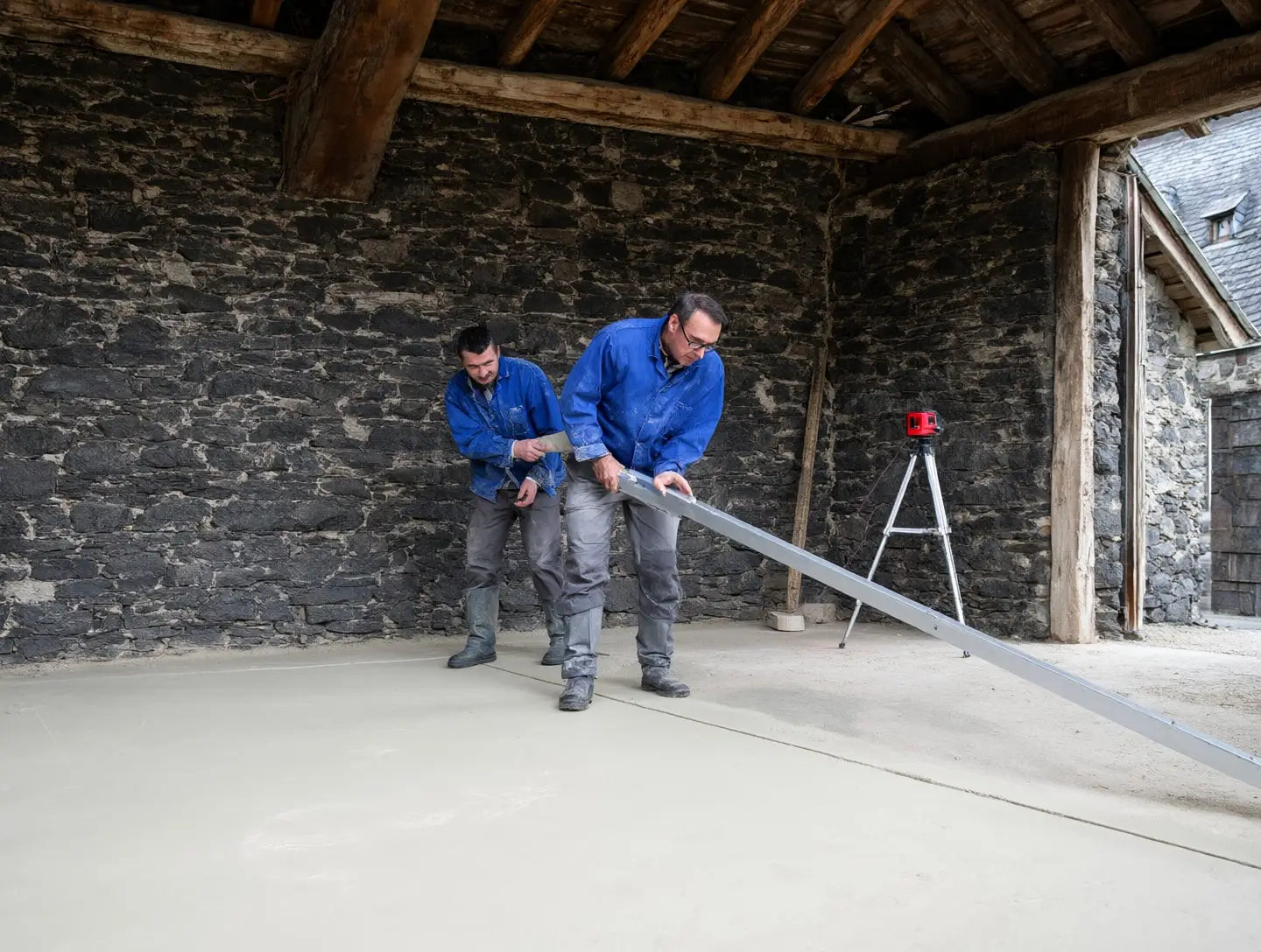 Deux artisans tirent à la règle une dalle de chaux fraîchement coulée dans une grange en pierre.