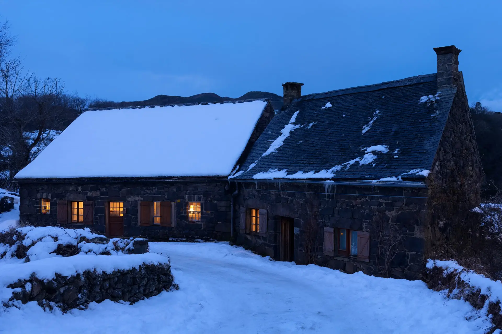 Deux maisons de pierre volcanique côte à côte dans un village enneigé du Cantal, à la tombée du jour. Le toit de gauche est entièrement couvert de neige, celui de droite l’a presque toute perdue.