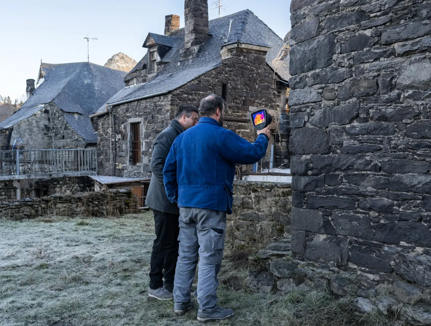 Un artisan en veste bleue pointe une caméra thermique vers une façade en pierre, à côté de la propriétaire.